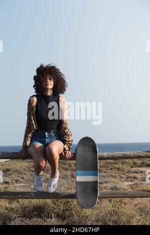 Jeune femme patineuse avec cheveux afro assis sur une clôture à l'extérieur regardant la caméra Banque D'Images