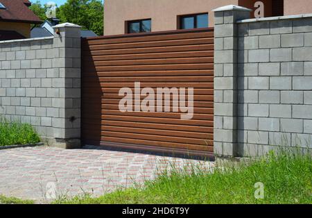 Un gros plan d'une barrière coulissante automatique à lattes de bois, d'une entrée, d'une clôture en brique grise avec piliers et d'une allée de finisseur devant une hutte Banque D'Images
