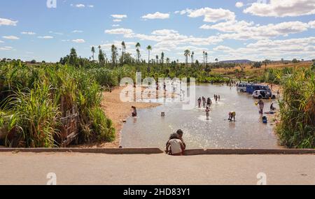 Ilakaka, Madagascar - 05 mai 2019 : groupe de malgaches lavant leurs vêtements et leurs voitures en rivière par la route, palmiers verts et buissons à proximité.Blanchisserie Banque D'Images