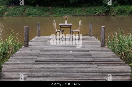 Coin reposant au bord de la rivière et fleurs séchées dans un vase blanc pour la décoration sur une table en bois ancienne et deux chaises en bois donnant sur la rive Banque D'Images