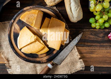 Vue aérienne d'un morceau de fromage provolone coupé sur une table et quelques raisins en arrière-plan. Banque D'Images