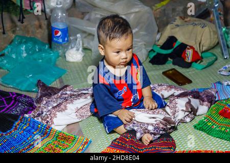 Un adorable bébé Chubby local se trouve dans une cabine de vente de tissus dans le marché nocturne de la rue piétonne dans le centre de Luang Prabang, le nord du Laos, l'Asie du sud-est Banque D'Images