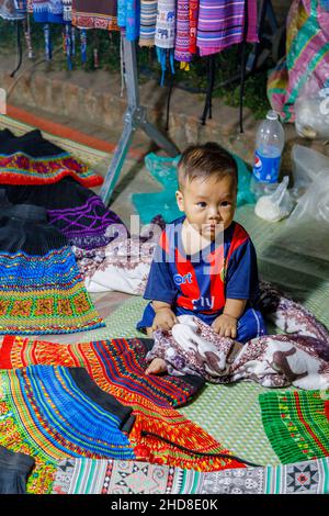 Un adorable bébé Chubby local se trouve dans une cabine de vente de tissus dans le marché nocturne de la rue piétonne dans le centre de Luang Prabang, le nord du Laos, l'Asie du sud-est Banque D'Images