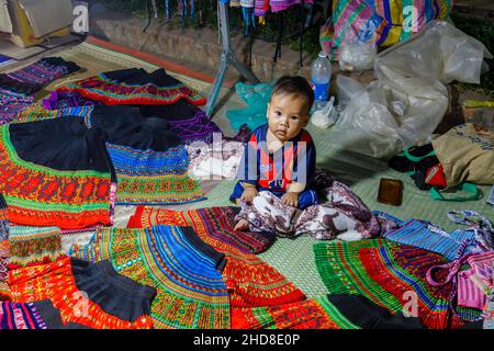 Un adorable bébé Chubby local se trouve dans une cabine de vente de tissus dans le marché nocturne de la rue piétonne dans le centre de Luang Prabang, le nord du Laos, l'Asie du sud-est Banque D'Images