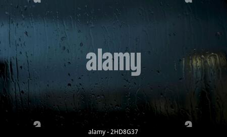 Des gouttes d'eau sur la fenêtre s'intarissent sur fond de ville tard en soirée.Ciel très nuageux et temps pluvieux, gros plan des fenêtres mouillées avec des gouttes d'eau. Banque D'Images