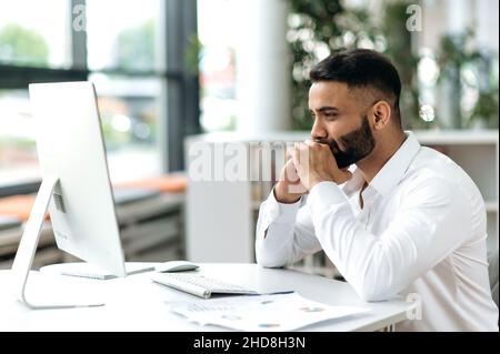 Sérieux et intelligent jeune homme d'affaires indien, directeur, indépendant avec barbe, assis dans un bureau à l'ordinateur, étudiant des documents, pensant à la stratégie d'affaires Banque D'Images