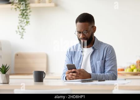 Un jeune homme afro-américain sérieux avec une barbe dans des lunettes, bavardant sur un smartphone, travaille à distance Banque D'Images