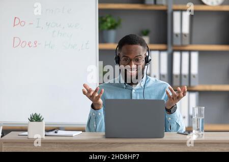 Joyeux jeune africain américain tuteur ou des gestes d'enseignant avec les mains et regarde l'écran de pc dans la maison Banque D'Images