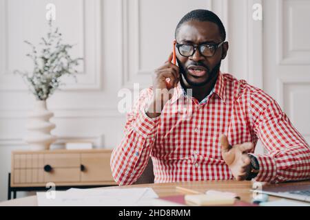 Homme d'affaires afro-américain insatisfait parlant émotionnellement sur un smartphone lorsqu'il est assis à son bureau Banque D'Images