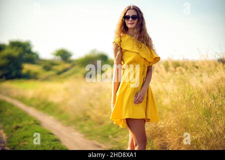 Belle fille en forme de curly marche sur le terrain en été tout en portant des lunettes de soleil et une robe jaune.Style de vie Banque D'Images