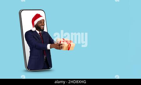 Homme d'affaires noir à chapeau de père Noël avec cadeau Peeking de Big smartphone Banque D'Images