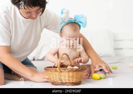 Maman et bébé avec des oreilles de lapin, avec des œufs de Pâques dans leurs mains, assis sur le canapé, parents et enfants jouent à l'intérieur. La famille fête Pâques Banque D'Images