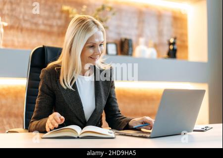 Amical heureux confiant intelligent influent mature femme d'affaires caucasienne, pdg ou expert financier travaillant dans un bureau moderne, en utilisant un ordinateur portable, en consultant des webinaires en ligne ou en communiquant par vidéo, en prenant des notes Banque D'Images