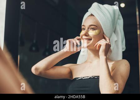 Gros plan portrait d'une fille à cheveux rouges avec une serviette sur la tête en appliquant des taches oculaires.Femme caucasienne souriante.Soin du visage.Nettoyer la peau.Beauté Banque D'Images