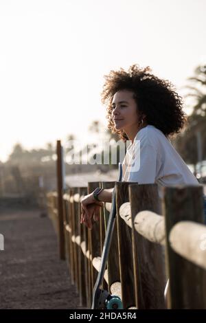 Skater jeune femme pensive penchée sur une clôture au coucher du soleil Banque D'Images