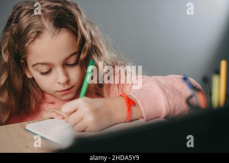 Gros plan d'une petite fille assise à une table et écrivant dans un carnet faisant ses devoirs, en se penchant sur le visage de sa main.Vue avant.Enfants Banque D'Images