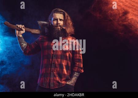 Le beau homme tient la vieille hache.Bûcheron barbu.Homme brutal et sérieux en chemise à carreaux aux cheveux longs Banque D'Images