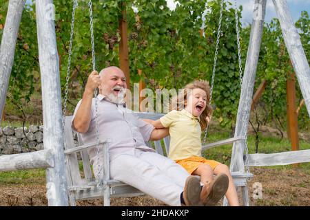 Jeune petit-fils et grand-père balançant dans le jardin de printemps.Grand-père et petit-fils assis sur le balançoire dans le parc. Banque D'Images