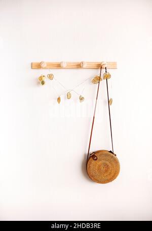 Image minimaliste d'un sac à bandoulière rond en rotin et boho suspendu sur un support en bois contre un mur blanc.Concept d'accessoires de mode et de style bohème.Min Banque D'Images