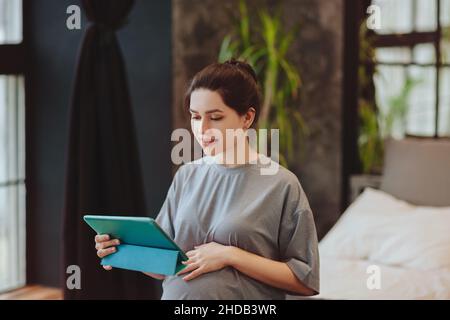 Jeune femme enceinte souriante tenant une table numérique et effectuant un appel vidéo tout en étant assise sur le lit à la maison, parlant en ligne avec sa famille ou ses amis.Distance p Banque D'Images