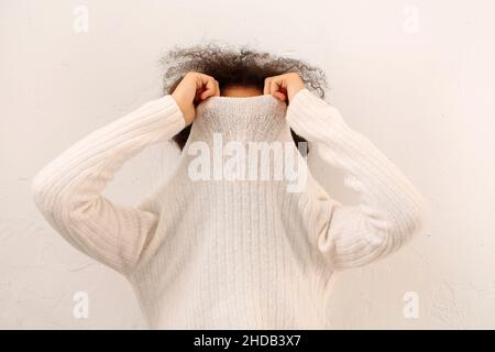 Une femme en forme de curly africaine couvre la face avec un col chandail blanc chaud isolé sur fond studio léger, une femme qui prend le pull à col roulé, ne peut pas chauffer Banque D'Images