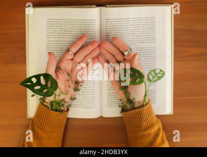 Feuilles vertes enveloppées autour des mains des femmes allongé sur un livre ouvert.Coupe courte de femme assise avec des paumes ouvertes à la table avec une plante qui pousse sous le sweat Banque D'Images