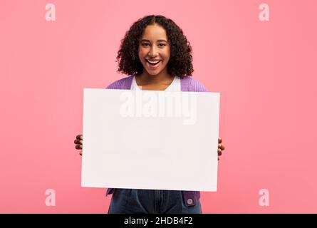 Bonne femme noire millénaire tenant une affiche vide, souriant à l'appareil photo sur fond rose studio, maquette Banque D'Images