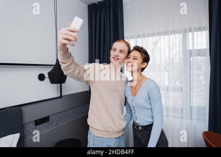 Un couple souriant prend son selfie sur son smartphone dans une chambre d'hôtel moderne Banque D'Images