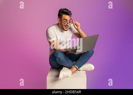 Homme arabe millénaire assis à pieds croisés avec un pc portable, touchant des lunettes, regardant l'écran en choc, lumière néon Banque D'Images