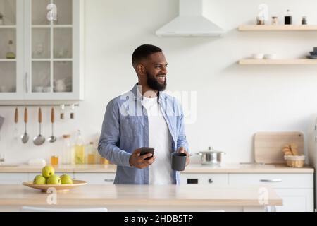 Un homme afro-américain satisfait avec la barbe aime le matin et boire dans la tasse avec un smartphone Banque D'Images