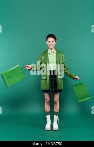 vue sur toute la longueur de la femme souriante dans une veste en cuir, une jupe à carreaux et des bottes blanches posant avec des sacs de shopping sur le vert Banque D'Images