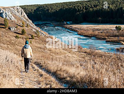 vue arrière de la femme aux couleurs neutres vêtements marchant par la rivière en automne sur l'herbe sèche contre les rochers et la forêt, tons de terre paysage, randonnée appréciant Banque D'Images