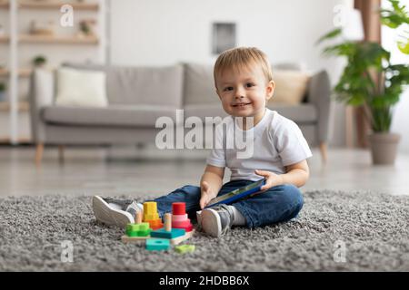Un petit garçon heureux regardant un dessin animé sur un téléphone intelligent tout en étant assis sur la moquette, en regardant et en souriant l'appareil photo Banque D'Images