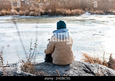 Jeune femme de derrière assise sur la pierre enembrassant chien regardant la rivière couverte de la première glace en automne ou au début de l'hiver, beige Colo neutre Banque D'Images