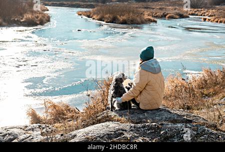 Jeune femme de derrière assise sur la pierre enembrassant chien regardant la rivière couverte de la première glace en automne ou au début de l'hiver, beige Colo neutre Banque D'Images