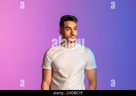 Portrait d'un jeune homme arabe sérieux en t-shirt blanc posant et regardant loin dans la lumière du néon Banque D'Images