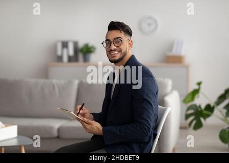 Portrait de psychothérapeute arabe masculin avec presse-papiers regardant et souriant à la caméra, travaillant au bureau moderne, espace de copie Banque D'Images