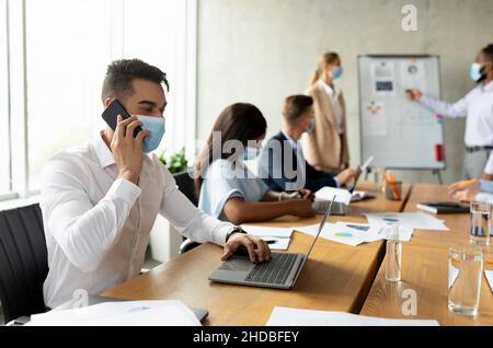 Masque de port d'un employé arabe utilisant un ordinateur portable et un téléphone portable lors d'une réunion avec des collègues Banque D'Images