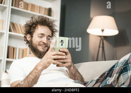 Guy fatigué se trouve dans son lit et joue Relax Game sur son smartphone avant le coucher.Un étudiant épuisé lit les nouvelles sur son mobile la nuit.Décor intérieur arrière-plan.Gros plan.Photo de haute qualité Banque D'Images