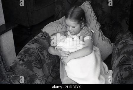 1950s, historique, une petite fille assise sur un canapé-lit tenant de près son bébé frère, un sommeil, enveloppé dans une couverture en laine, Angleterre, Royaume-Uni. Banque D'Images