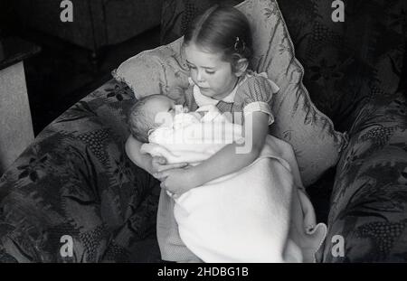 1950s, historique, une petite fille assise sur un canapé-lit tenant son bébé frère, enveloppé dans une couverture en laine, qui a ses yeux ouverts, Angleterre, Royaume-Uni. Banque D'Images