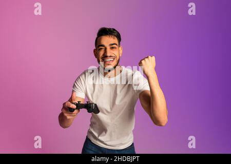 Homme arabe millénaire excité avec joystick célébrant la victoire du jeu vidéo, en gestant OUI dans la lumière de néon Banque D'Images