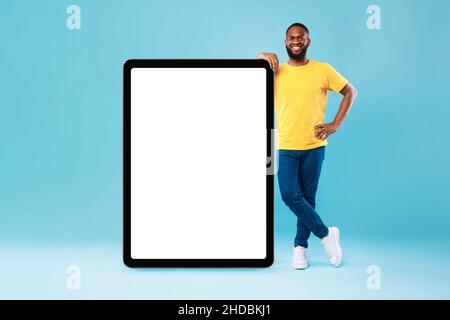 Joyeux jeune homme noir se penchant sur un PC tablette géant, démontrant écran blanc sur fond bleu, maquette Banque D'Images