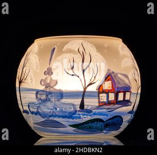 Une tasse en verre peinte à la main avec un décor hivernal éclairé par une bougie à l'intérieur de la tasse.Un gros plan photographique utilisant la peinture légère. Banque D'Images