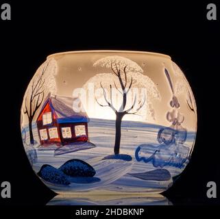 Une tasse en verre peinte à la main avec un décor hivernal éclairé par une bougie à l'intérieur de la tasse.Un gros plan photographique utilisant la peinture légère. Banque D'Images