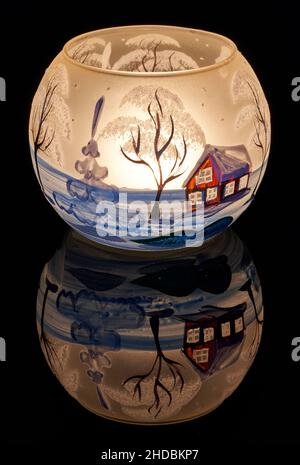 Une tasse en verre peinte à la main avec un décor hivernal éclairé par une bougie à l'intérieur de la tasse.Un gros plan photographique utilisant la peinture légère. Banque D'Images