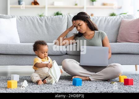 Femme noire stressée essayant de travailler sur un ordinateur portable avec un petit bébé autour Banque D'Images