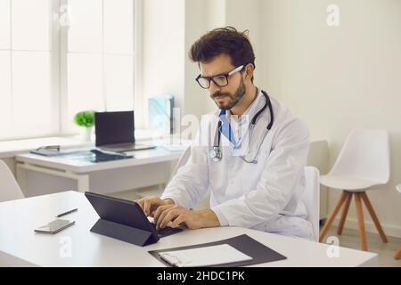 Un jeune médecin sérieux est assis au bureau et travaille sur une tablette Banque D'Images