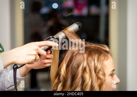 Coiffeur fait des boucles avec un fer à friser pour la jeune femme avec de longs cheveux bruns dans un salon de beauté Banque D'Images