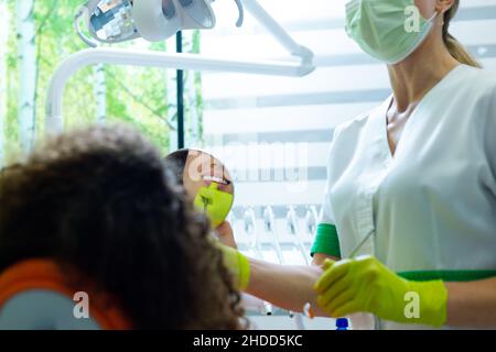 Belle adolescente vérifiant ses dents dans le miroir après traitement dentaire, femme dentiste debout.Vue sur l'épaule du patient. Banque D'Images